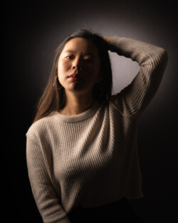 A portrait of a woman in a textured sweater posing against a dark background with dramatic lighting highlighting one side of her face. She has her arm raised, hand behind her head, and wears hoop earrings. Her expression is serene and introspective, creating a moody and artistic photograph.