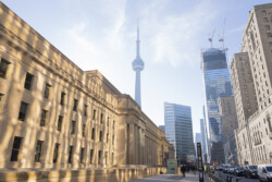 A cityscape showcasing the contrast between historical and modern architecture. On the left, a classical stone building basks in the warm sunlight, while on the right, contemporary glass skyscrapers rise towards the sky. In the distance, the iconic CN Tower dominates the skyline, symbolizing the city of Toronto. The scene is a busy street with pedestrians and traffic, indicative of a vibrant urban center.