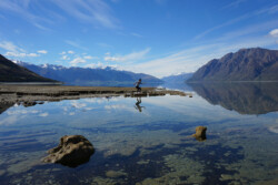 A panoramic view of a serene lake with a mirror-like reflection of the surrounding mountains. The mountains have snowy peaks and the sky is partly cloudy. In the foreground, a person is skipping across a narrow strip of land that extends into the water, creating a playful interaction with their reflection on the water's surface.
