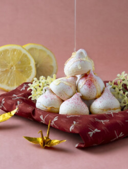 The image displays a pile of white meringues with red sprinkles on a red patterned cloth, with lemon slices and two origami cranes beside them, against a pink background. A photograph showcases a stack of lemon meringue kisses on a red cloth with a delicate white floral pattern. A glistening stream of honey is being drizzled over the top meringue, accentuating its white peaks with a golden sheen. To the left, two slices of fresh lemon provide a bright contrast, while in the foreground, an intricate golden origami crane adds an artistic touch to the composition set against a soft pink background.