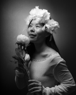 A black and white portrait of a smiling woman looking upwards, wearing a flower crown made of white blooms. She is holding a bouquet of similar white flowers, exuding a sense of joy and whimsy. The soft lighting and grayscale tones give the image a timeless, ethereal quality.