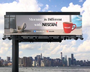 An outdoor billboard advertisement featuring NESCAFÉ, positioned against a city skyline viewed from across a body of water. The billboard showcases a large red NESCAFÉ mug filled with coffee on the right, with the tagline "Morning is Different with NESCAFÉ." On the left side, there's a shattered white coffee pot and a cup, implying a contrast between traditional coffee-making methods and the convenience of NESCAFÉ. Social media icons and the website 'Nescafe.CA' are displayed at the bottom, inviting viewers to connect online. The scene is set under a blue sky with fluffy clouds, and there are birds flying over the water, adding to the urban morning ambiance.