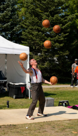 A performer, dressed in a white sleeveless shirt with red suspenders and striped pants, juggles multiple leather-wrapped balls in the air. He is looking up at the balls with an expression of concentration and delight. The setting is outdoors with a tent and trees in the background, suggesting a festival or outdoor event where the entertainer is showcasing his skills to an audience.