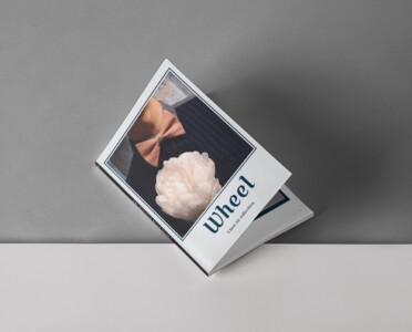 The image features a magazine resting on a flat surface with a grey background. The cover of the magazine displays the word "Wheel" in large, bold letters, indicating the title or the theme of the issue. Below the title, there is a subtitle that reads "Auto Tie Collection," suggesting that the magazine's content is related to automotive-themed neckwear. The cover photo showcases a close-up of a person wearing a bow tie, with a distinctive wheel-like design on the knot, reinforcing the automotive theme. A decorative white flower is placed on the magazine, adding an aesthetic touch to the presentation. The overall setting is minimalistic, drawing the viewer's attention to the magazine cover and its unique fashion content.