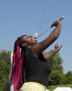 A performer with dark skin and striking red and white braided hair is engaged in a fire-eating act. She tilts her head back, mouth open, about to interact with a flaming baton held aloft in her hand. She wears a black top and yellow and white striped pants. The sky is clear and blue, highlighting the boldness of the act, with a hint of greenery in the distant background, suggesting an outdoor performance setting.