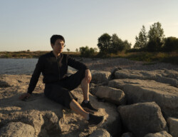 A man is sitting on some rocks at the lakeshore under the sunset. He appears to be looking at something far away. A young man with glasses is seated on rocky terrain near a body of water during the golden hour. He's dressed in a casual black zip-up sweater, black shorts, and sneakers. His pose is relaxed, one hand resting on the rock and the other on his raised knee. Trees and shrubbery can be seen in the background, creating a peaceful and natural setting.