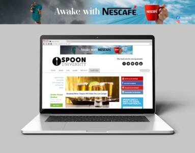 Copy space table with office supplies on workspace. A laptop screen displaying the homepage of "Spoon University," which is described as a food site for young people. The website features a top banner with the text "Awake with NESCAFÉ" alongside an image of a red NESCAFÉ mug. There are various navigation options including "COOK," "DRINK," "LIVE," "LEARN," "EAT OUT," and "CAMPUSES." Additionally, the site offers options to order food online from campus restaurants. Social media icons indicate the site's presence on platforms like Facebook, Twitter, YouTube, Pinterest, and Instagram. The laptop is set against a backdrop with a dark cloudy sky, and above the screen is an image of a hand holding a coffee bean with the NESCAFÉ logo, suggesting a promotional partnership or advertisement.