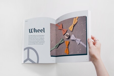 A person is holding a magazine open to a two-page feature about a brand called "Wheel." The left page has a large title "Wheel" and an introductory text about Jeffrey and Ian, two car-loving fashion designers known for their unique approach to bow ties. It mentions their journey from wearing old-school "weird" bow ties to founding "Wheel," a brand that offers distinctive bow ties and cufflinks. The right page shows a creatively arranged display of various bow ties in different colors and a pair of cufflinks on a textured background, signifying the quirky and innovative designs offered by the brand. The magazine spread is designed to provide readers with a story behind the brand as well as a visual representation of the products.