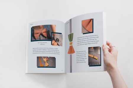 A person is holding open a magazine to a two-page article that features various bow ties and cufflinks. On the left page, there's a close-up image of a person wearing a clay-colored bow tie with a matching cufflink. The text below discusses the infrequent use of bow ties in contemporary fashion, noting that bow ties are often seen as antiquated but are being reintroduced by Jeffrey and Ian as a cool trend. Pricing for the bow tie and cufflink is listed at $90 each. On the right page, the text continues to promote a simplistic and practical fashion approach, suggesting organic cotton bow ties as a comfortable and allergy-free option. An image of a neatly folded bow tie with an organic label and another of a bow tie clip is shown, emphasizing ease of use. The magazine's layout is clean, with a focus on the products and their unique selling points.